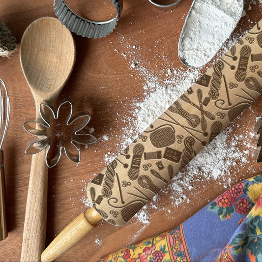 Wooden rolling pin with embossed designs on a wooden surface with flour and kitchen utensils.