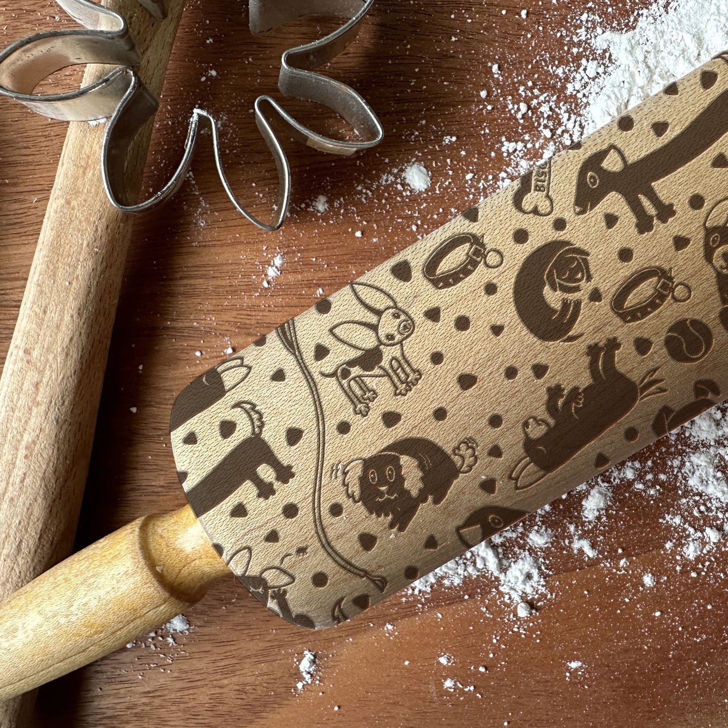 Rolling pin with embossed designs on a wooden surface with flour and cookie cutters.