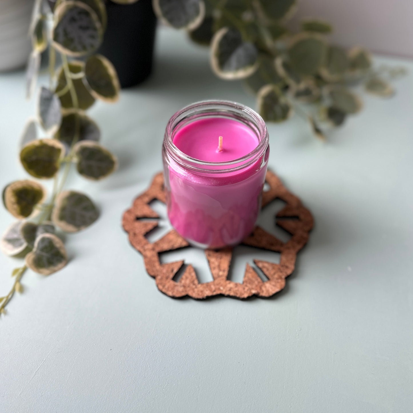 Pink candle in a glass jar on a decorative trivet with a plant in the background