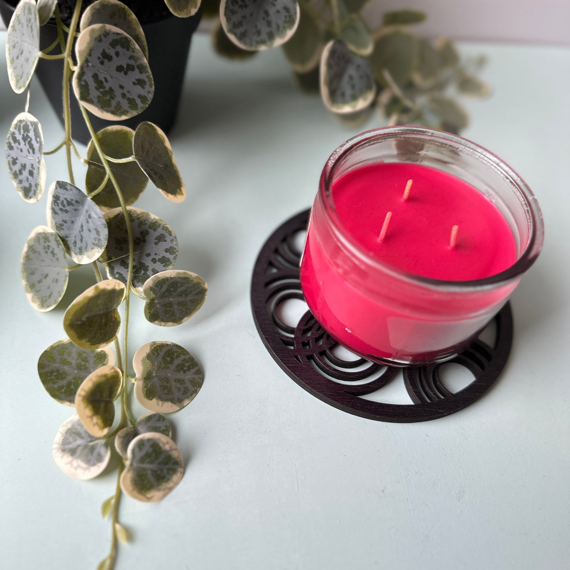 Pink candle in a glass holder on a black trivet with green leaves in the background