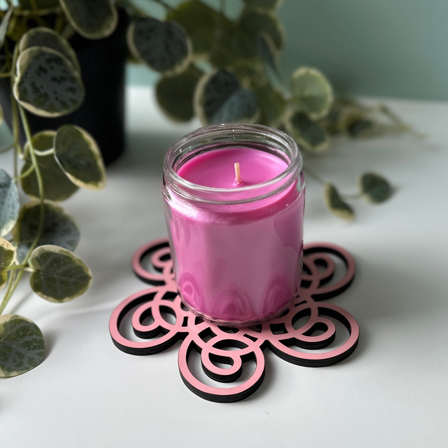 Pink candle in a glass jar on a decorative metal coaster with a plant in the background.