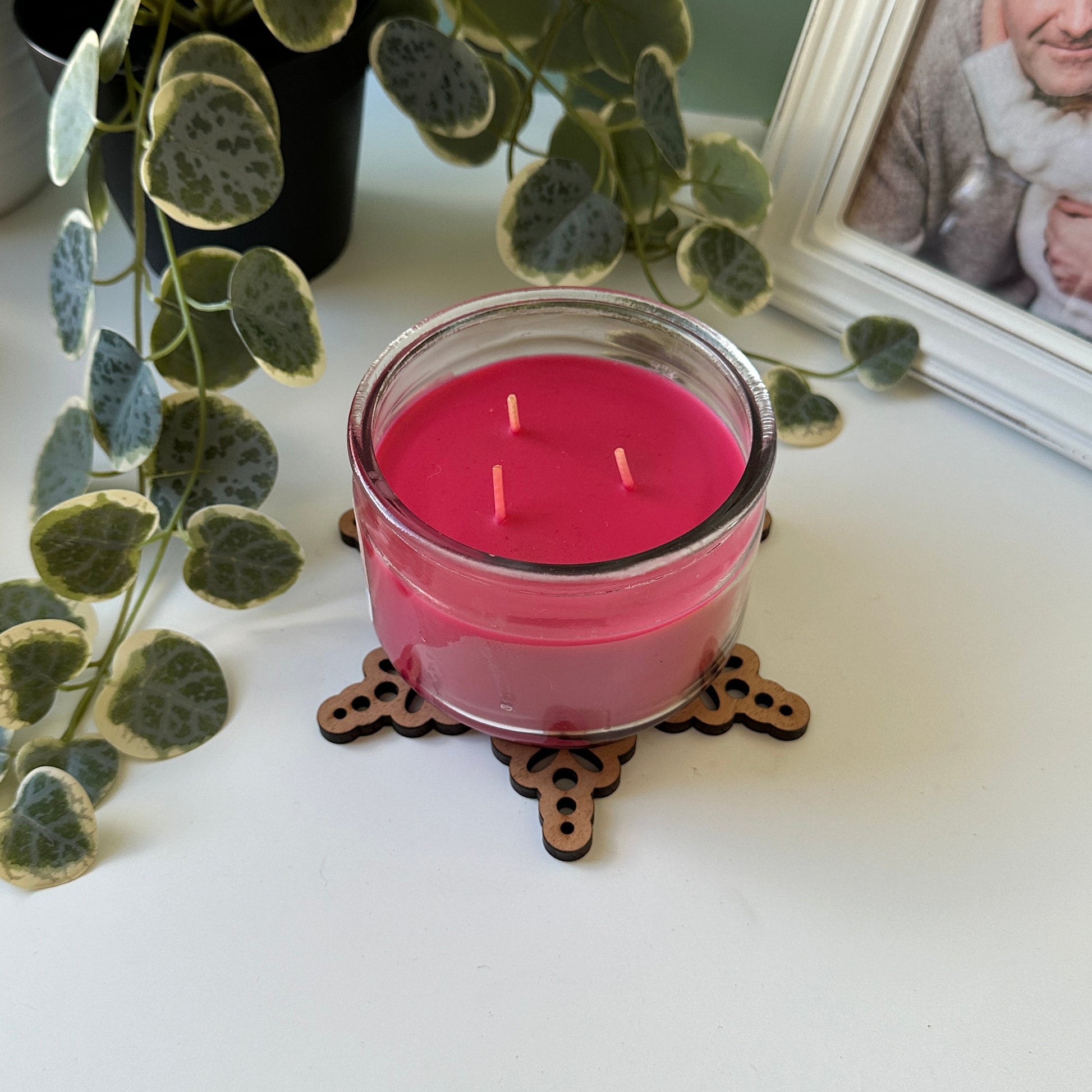 Pink candle on a decorative trivet  with a plant and photo frame in the background