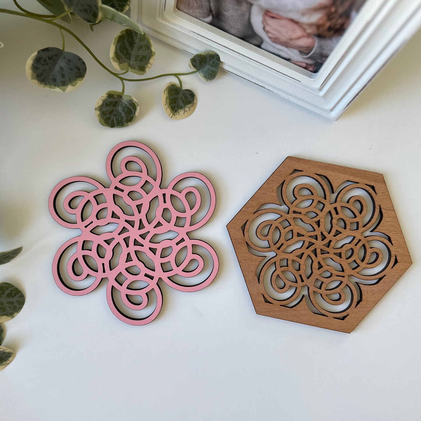 Pink and wooden geometric coasters on a white surface with green leaves.