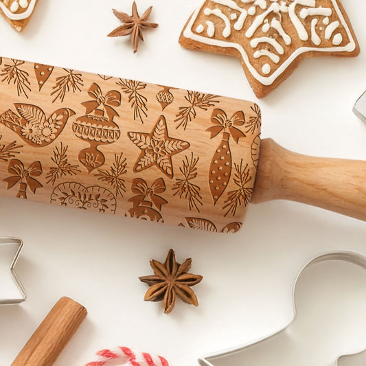 Wooden rolling pin with Christmas patterns surrounded by cookies, spices, and a spatula on a white surface.