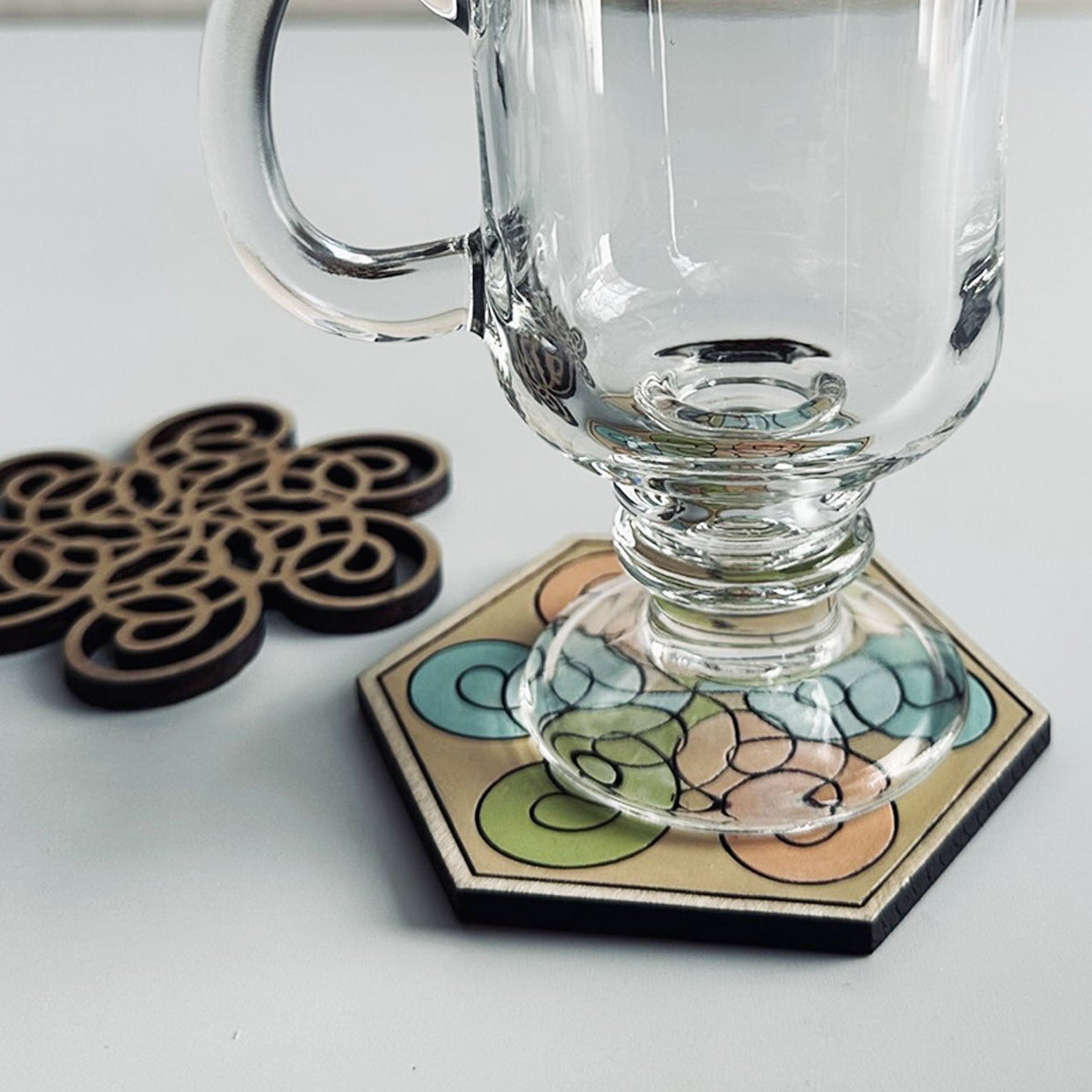Clear glass mug on a colorful hexagonal coaster with wooden coasters beside it on a white surface.