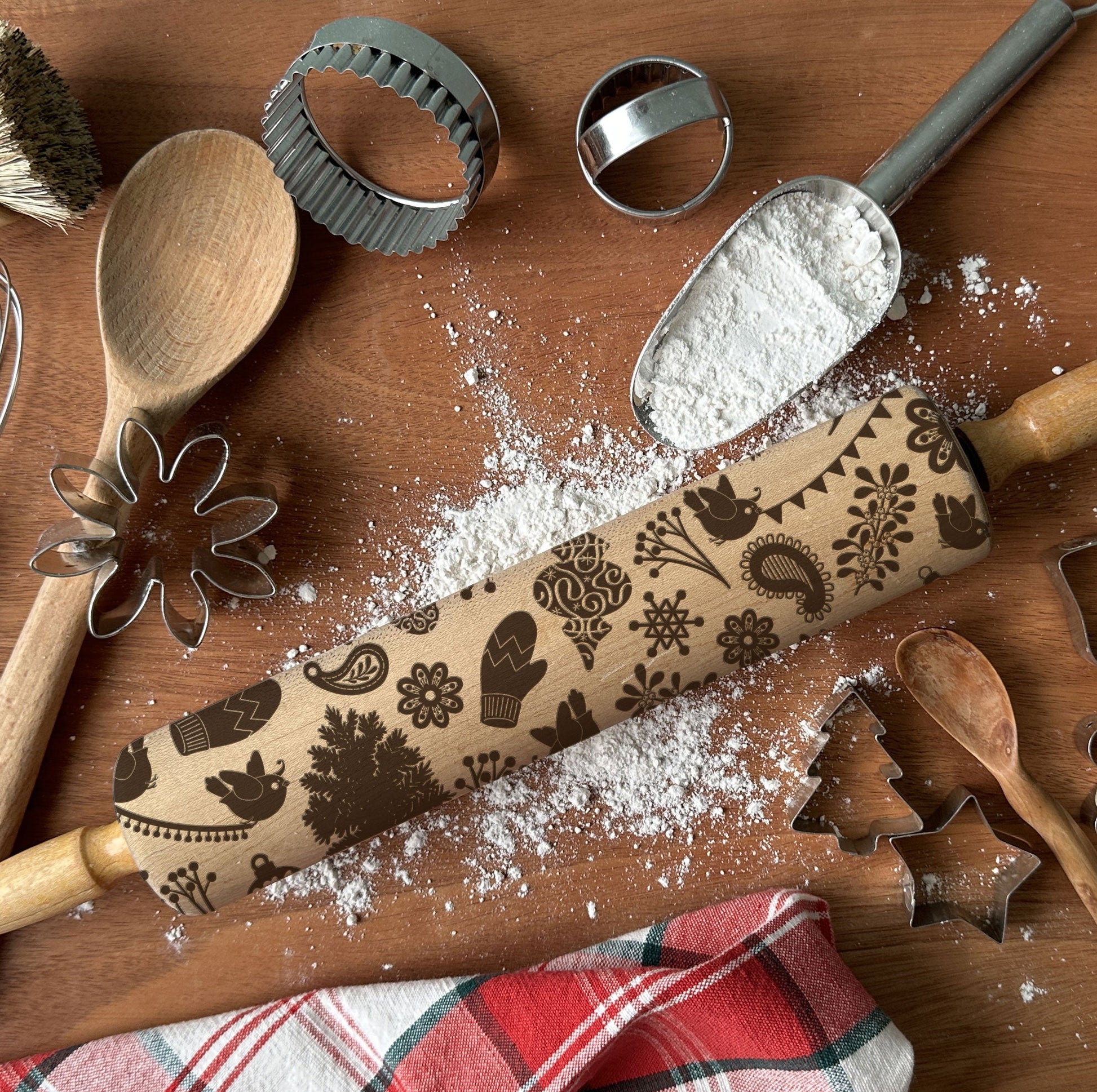 Baking tools including a rolling pin with embossed designs, wooden spoon, whisk, and cookie cutters on a wooden surface.