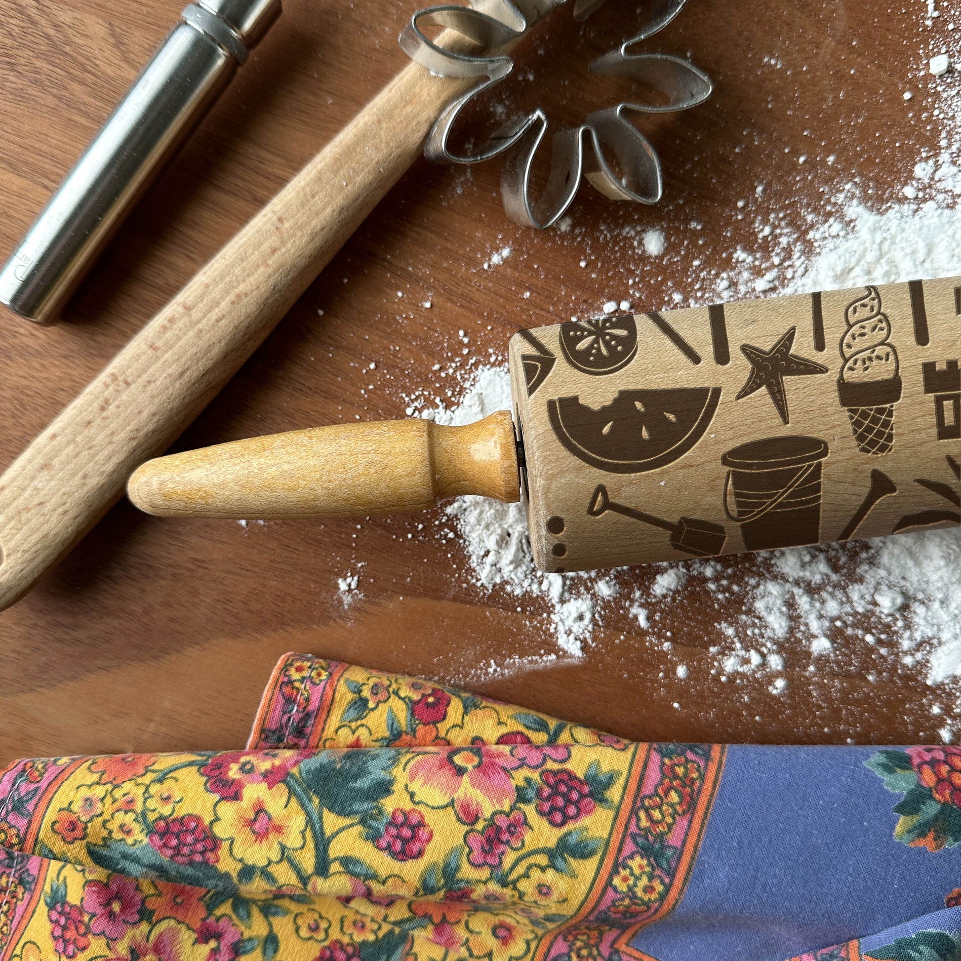 Wooden rolling pin with embossed designs on a wooden surface with flour and a colorful kitchen towel.