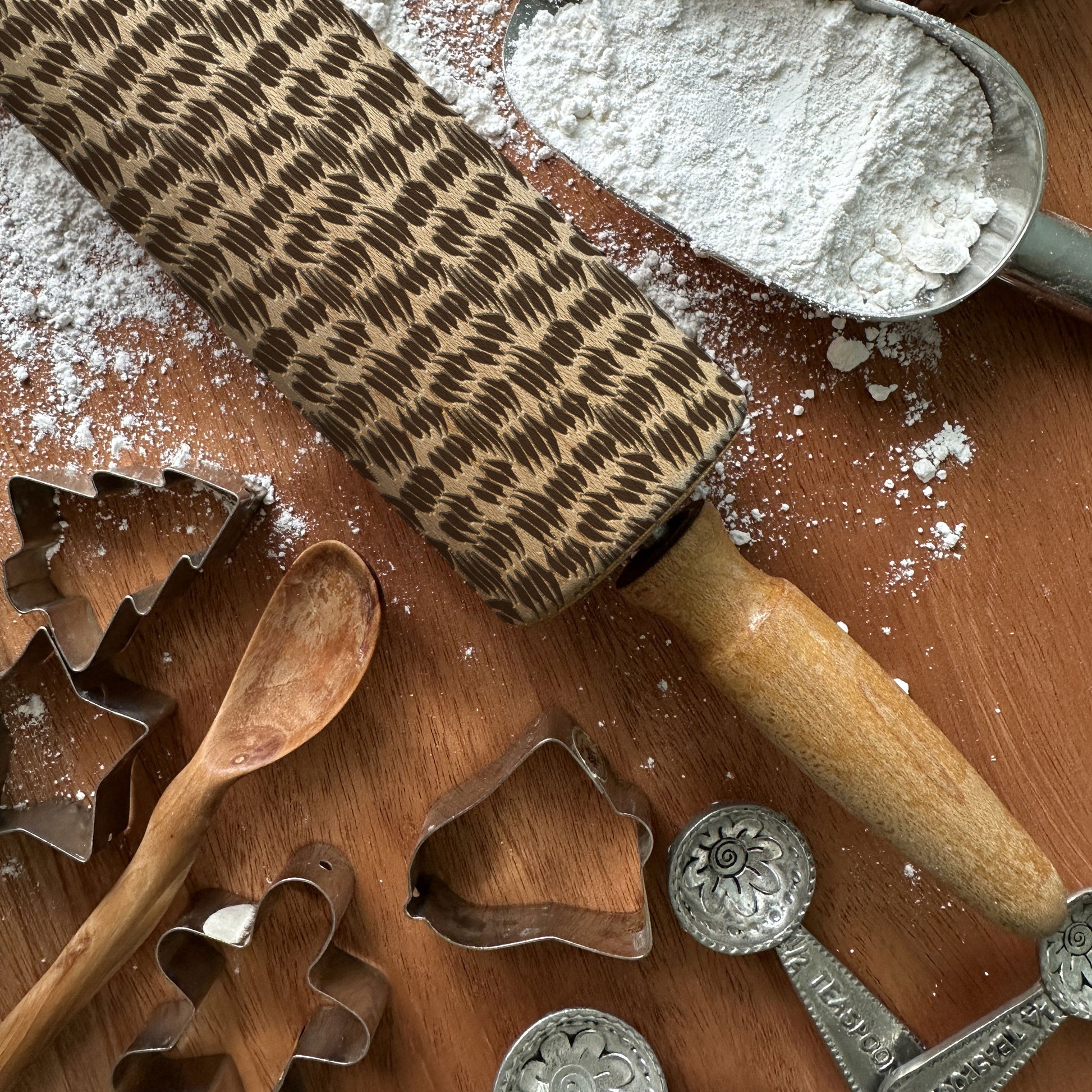 Wooden rolling pin with pattern, flour, wooden spoon, and metal cookie cutters on a wooden surface.