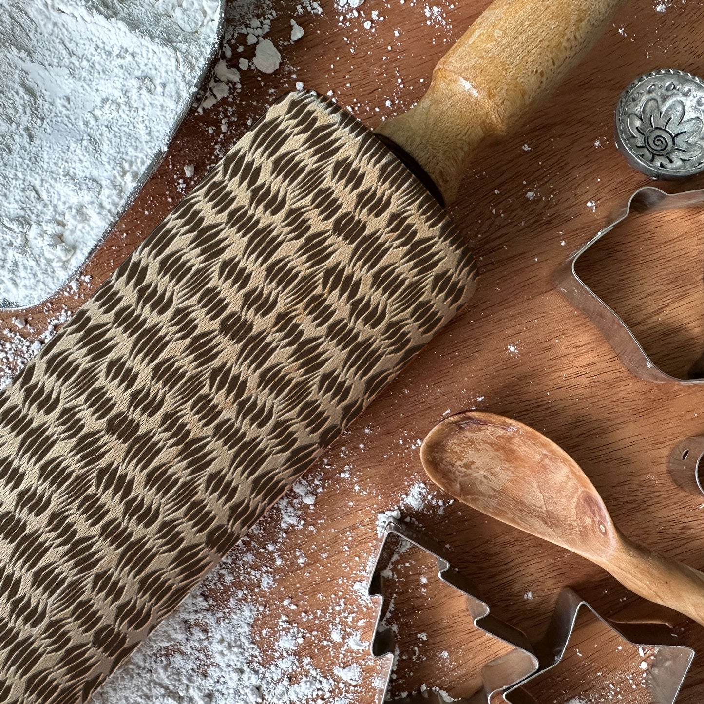 Wooden rolling pin with pattern, wooden spoon, and cookie cutters on a flour-dusted surface.