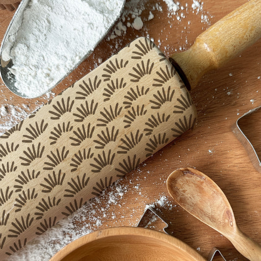 Rolling pin with pattern, cookie cutters, and wooden spoon on a wooden surface with flour, branded 'Pickle and Peach Studio'.