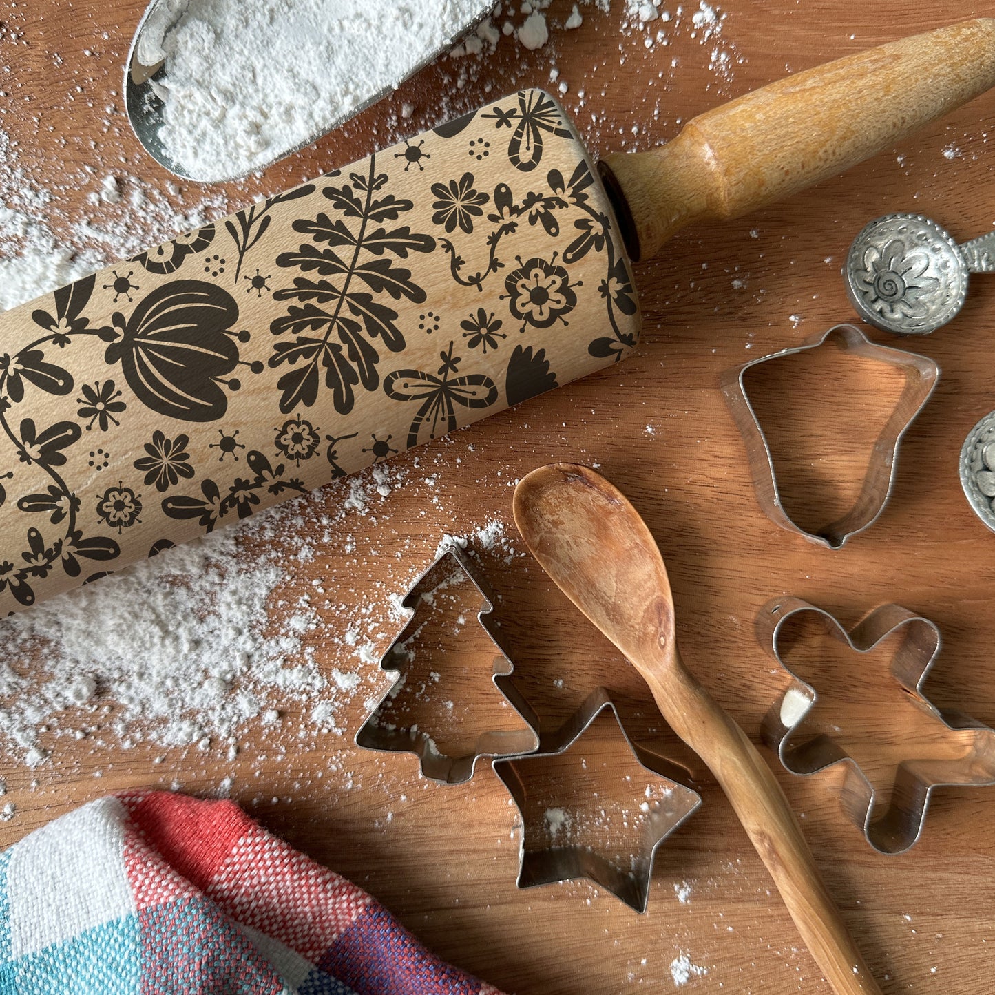 Baking tools including a floral-patterned rolling pin, cookie cutters, and a wooden spoon on a wooden surface with flour.