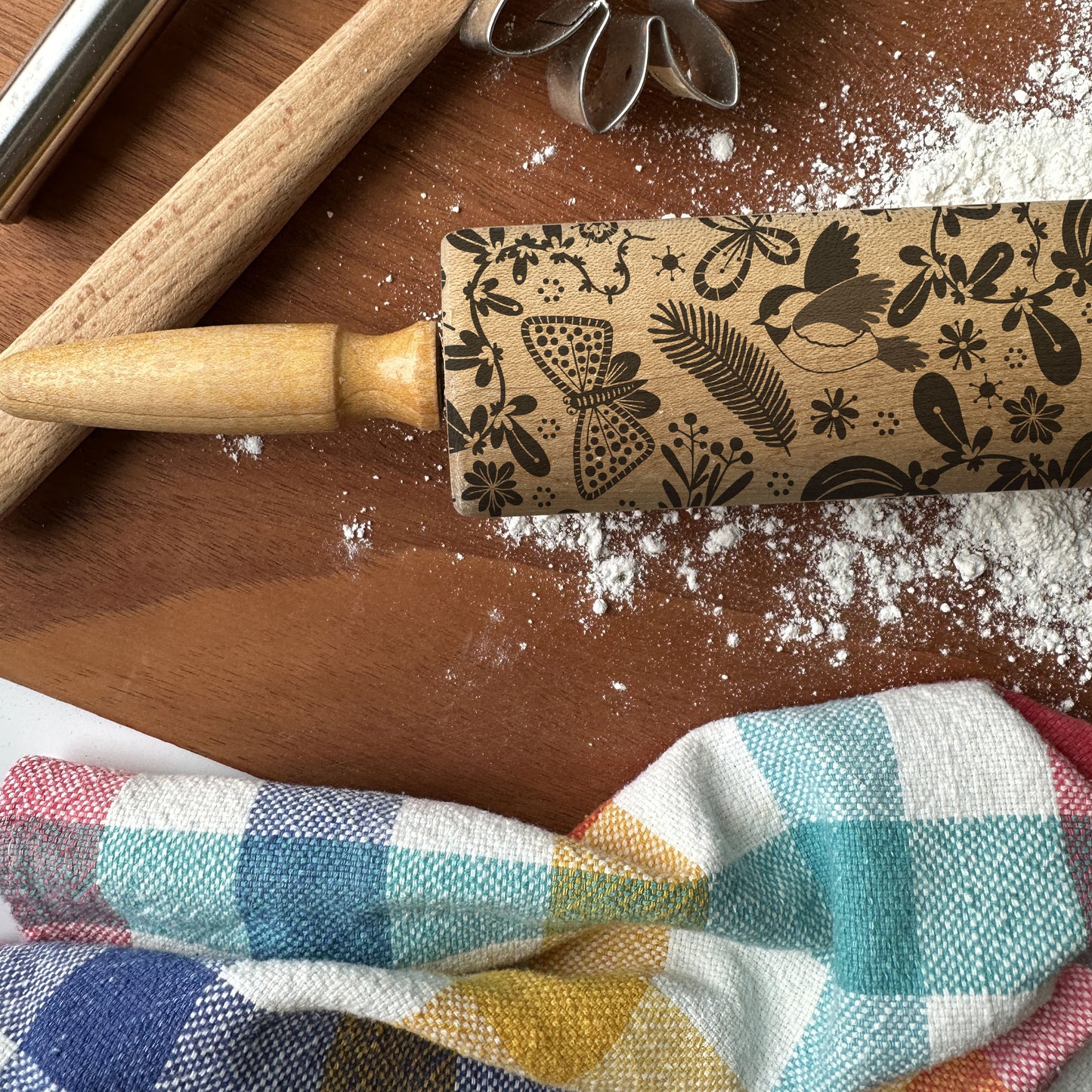 Decorative rolling pin with embossed patterns on a wooden surface with flour and a checkered cloth.