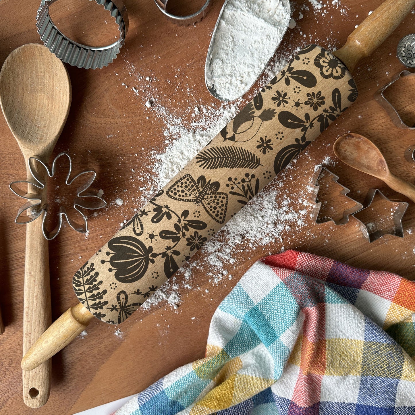 Baking tools including a rolling pin with floral designs, wooden spoon, whisk, and cookie cutters on a wooden surface with flour.