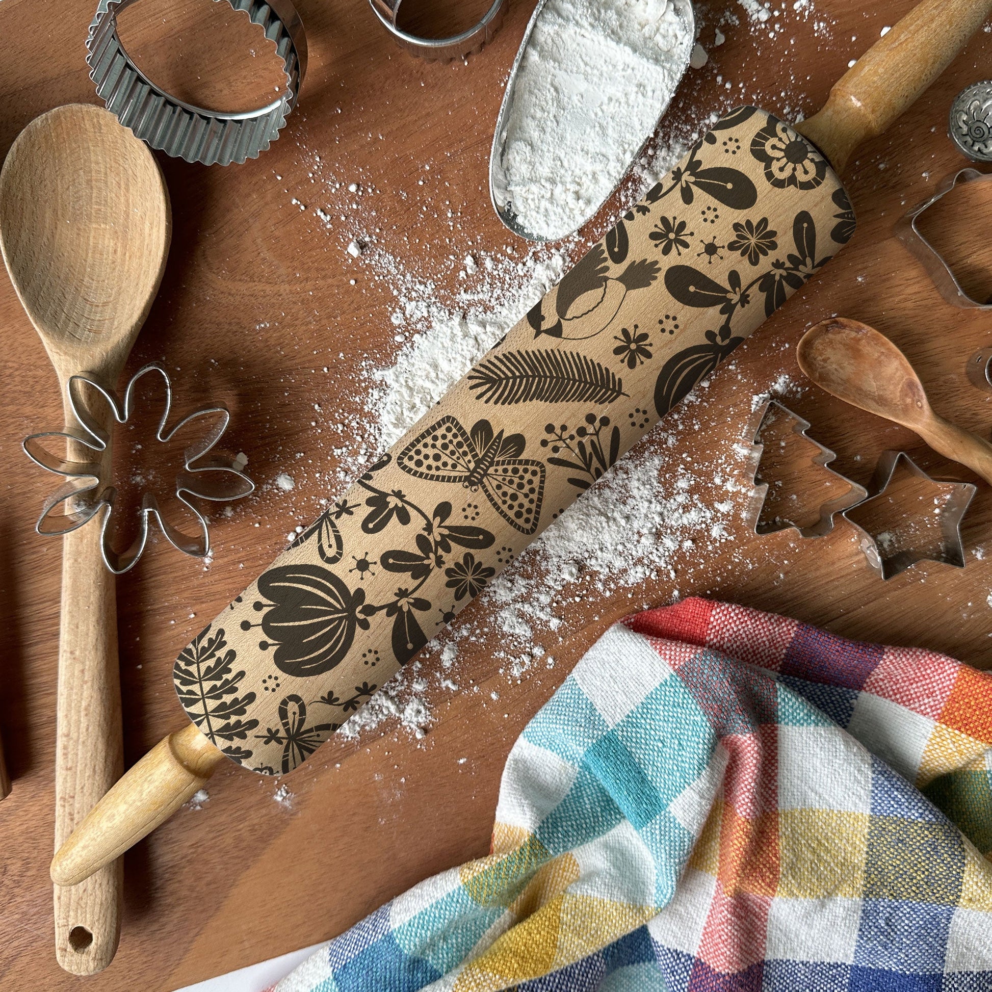 Baking tools including a rolling pin with floral designs, wooden spoon, whisk, and cookie cutters on a wooden surface with flour.