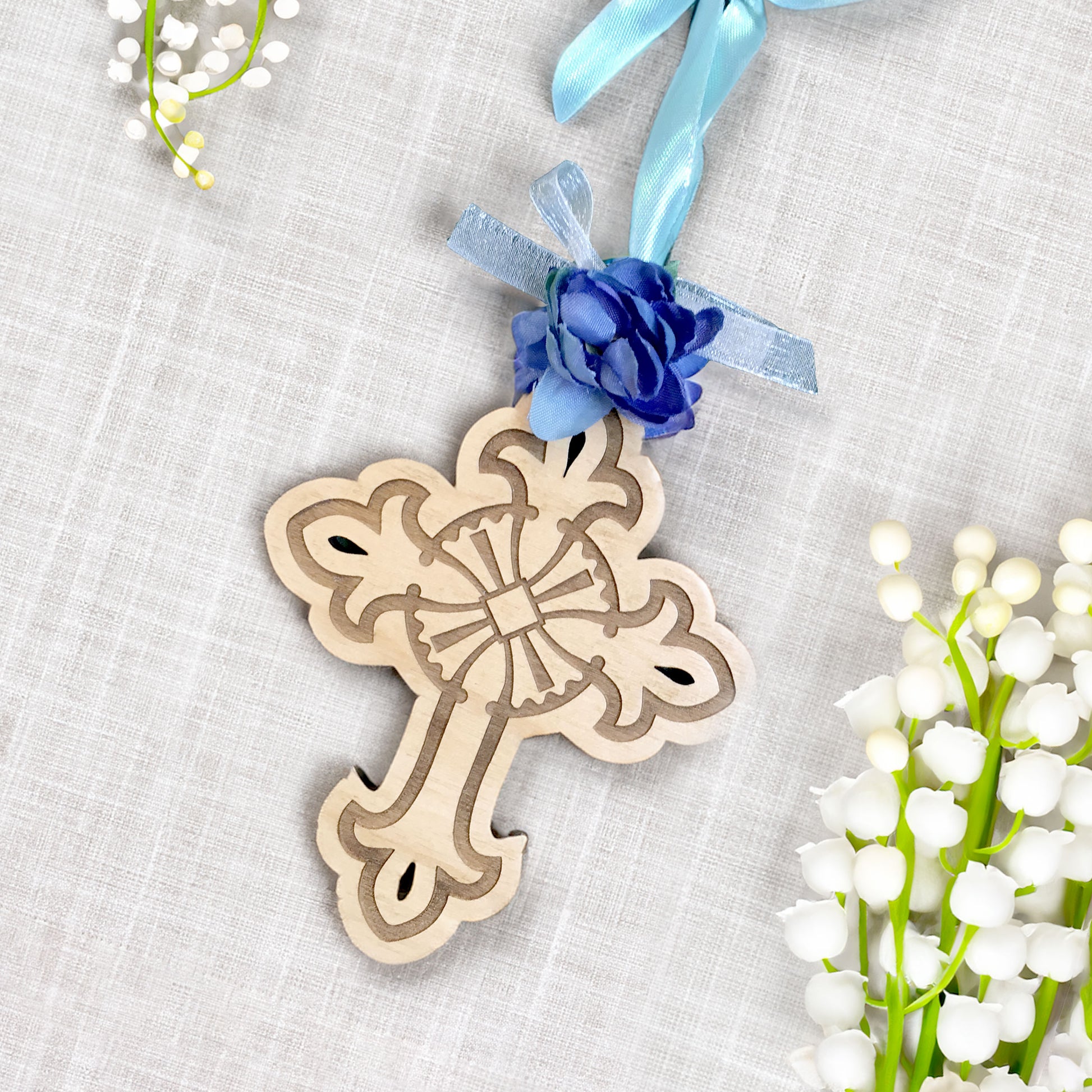 Wooden cross ornament with blue flower and ribbon on a textured surface with flowers.