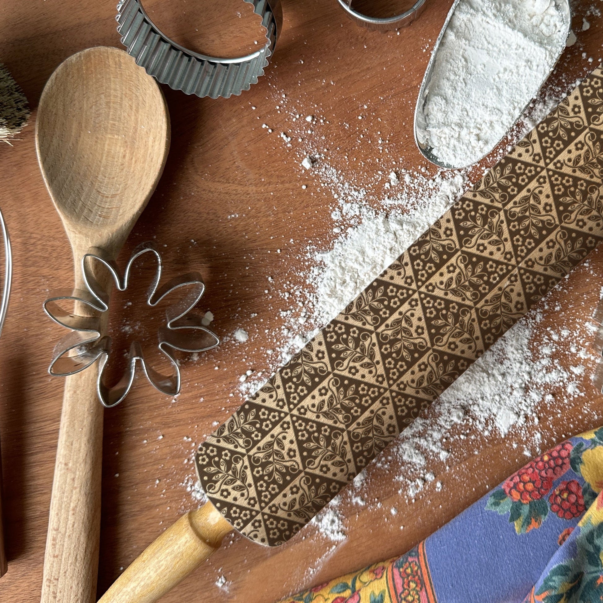 Baking tools on a wooden surface with flour and a colorful cloth underneath.