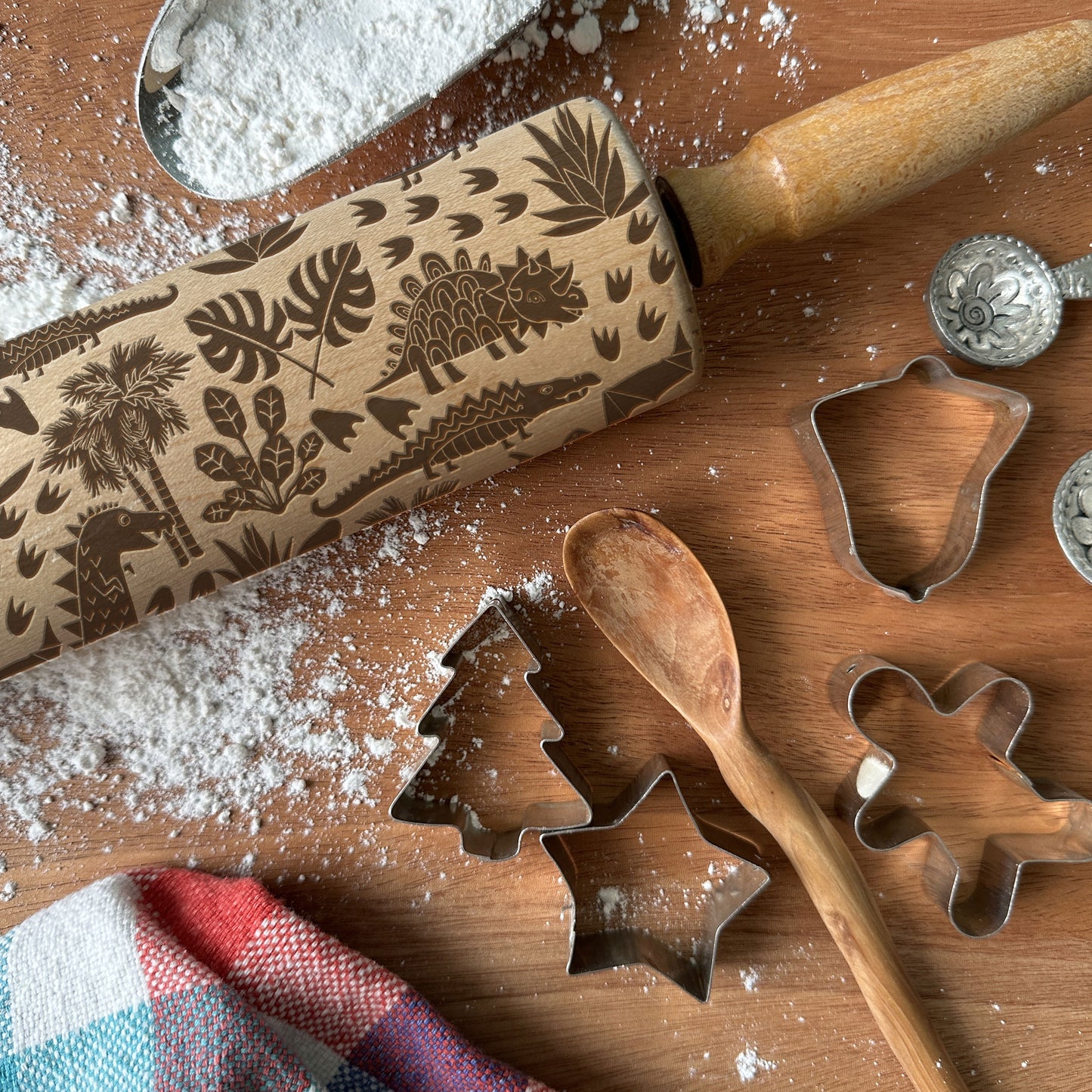 Baking tools including a rolling pin with dinosaur design, cookie cutters, and a wooden spoon on a wooden surface.