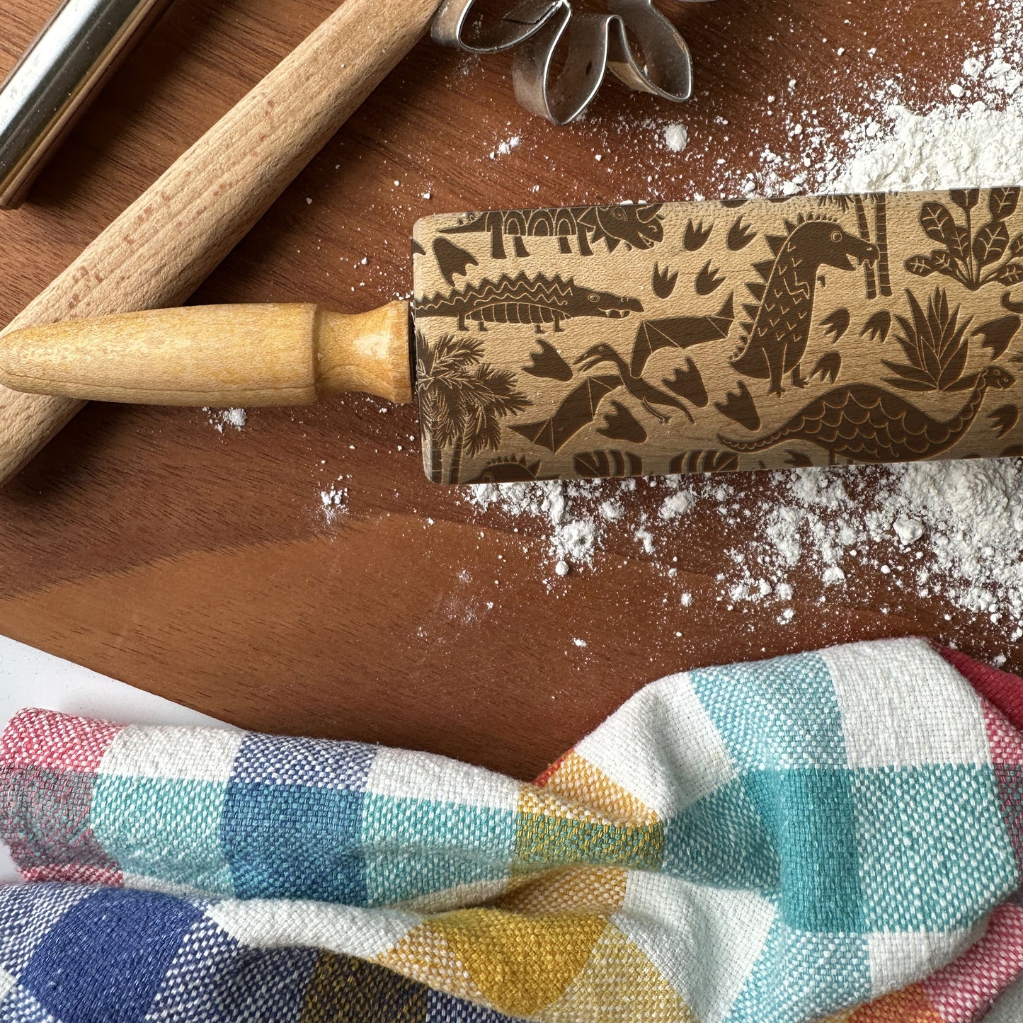 Wooden rolling pin with dinosaur pattern on a wooden surface with flour and checkered cloth.