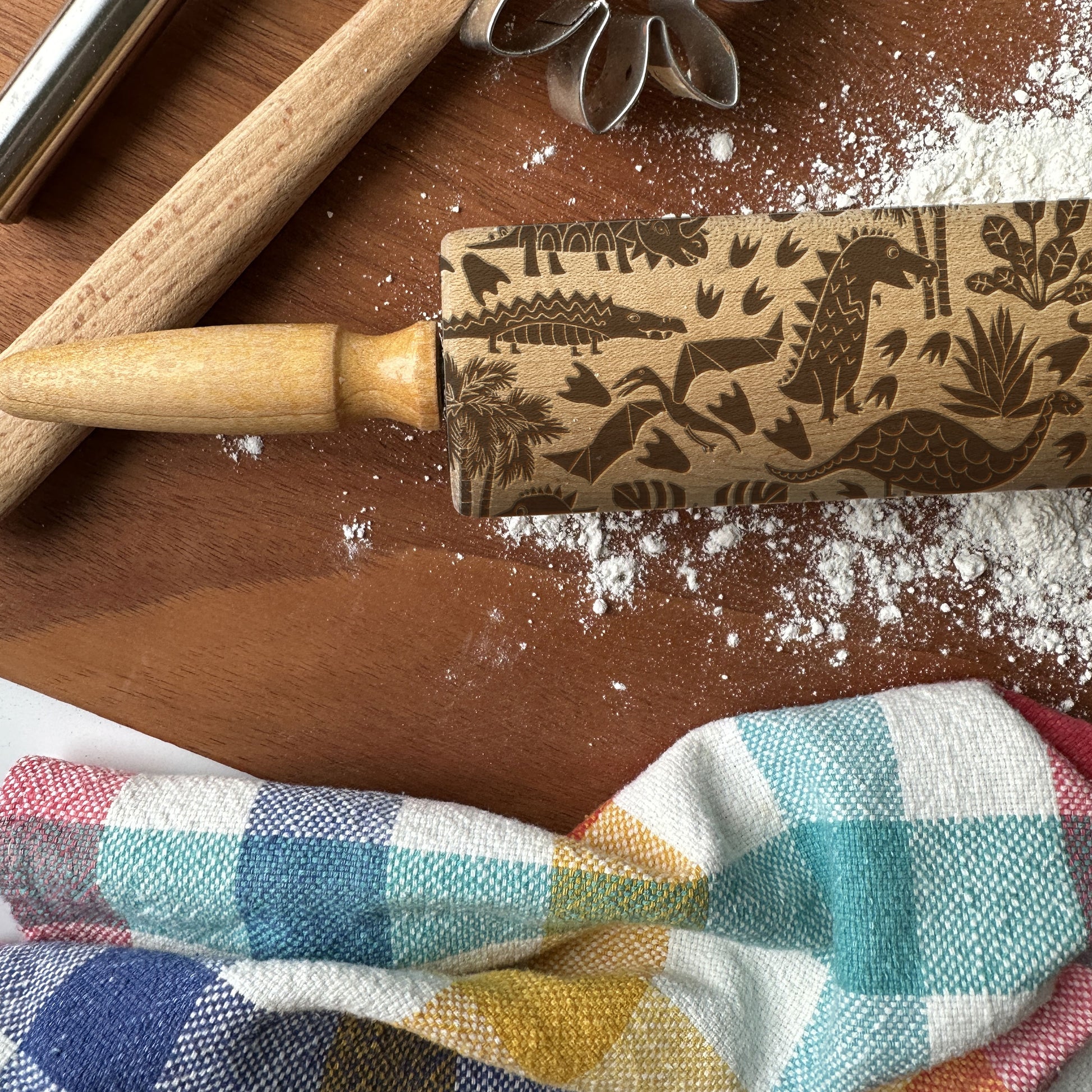Wooden rolling pin with dinosaur pattern on a wooden surface with flour and checkered cloth.