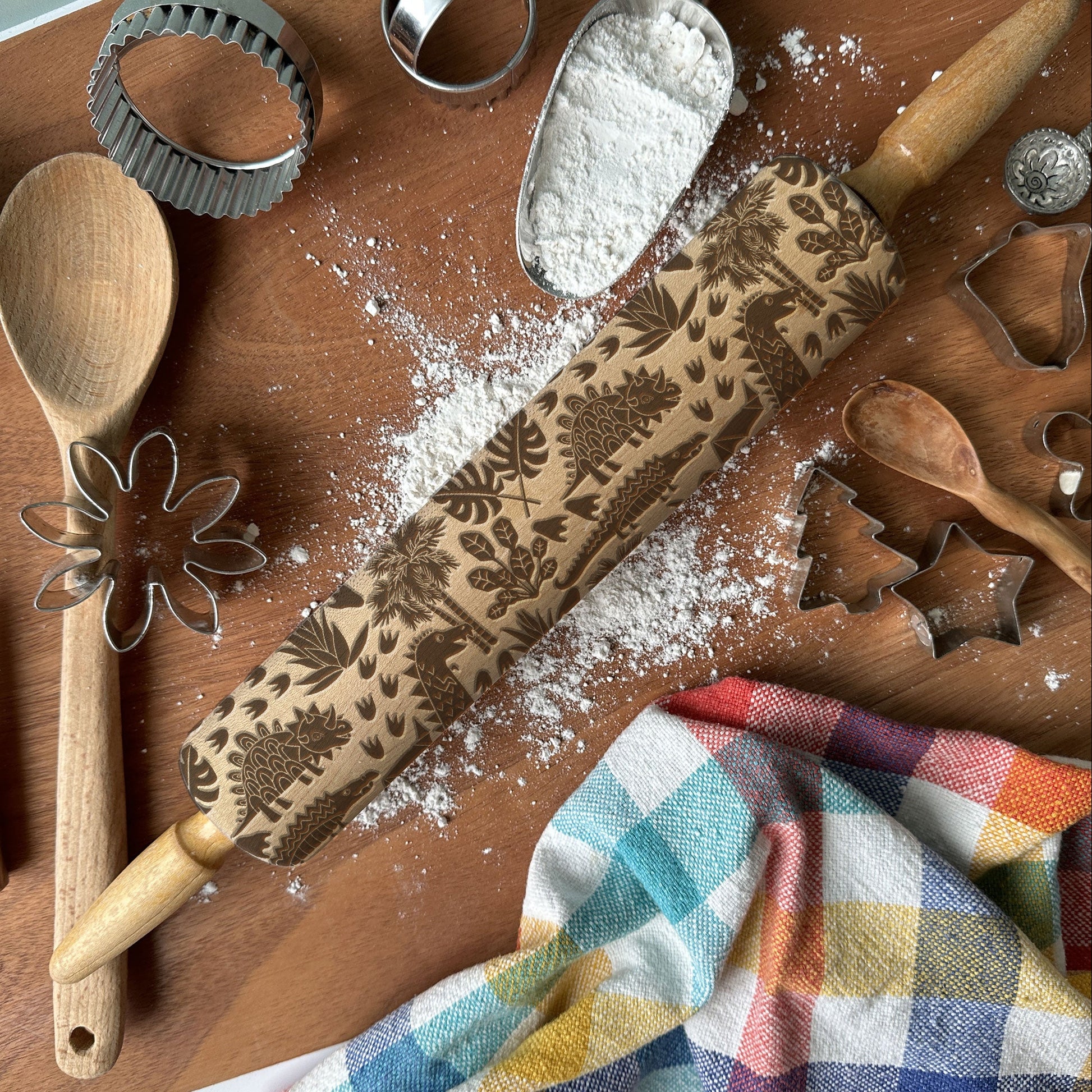 Baking tools including a rolling pin with floral patterns, wooden spoon, whisk, and flour on a wooden surface.