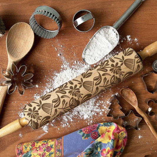Wooden rolling pin with floral patterns on a wooden surface with flour and baking tools.