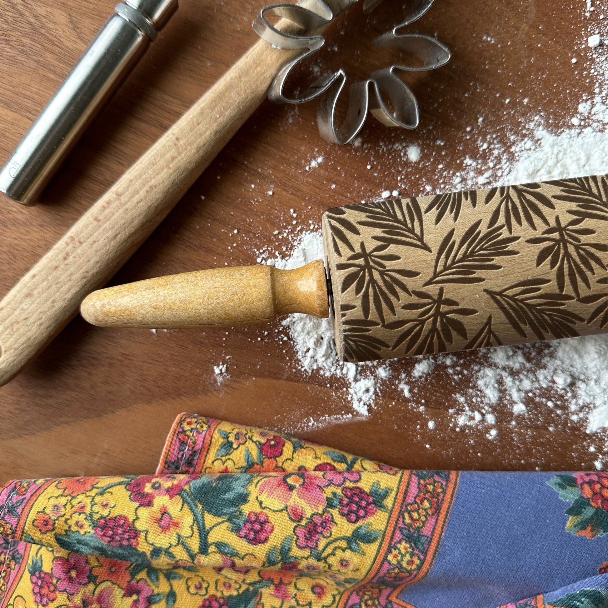 Decorative rolling pin with leaf pattern on a wooden surface with flour and a colorful towel.