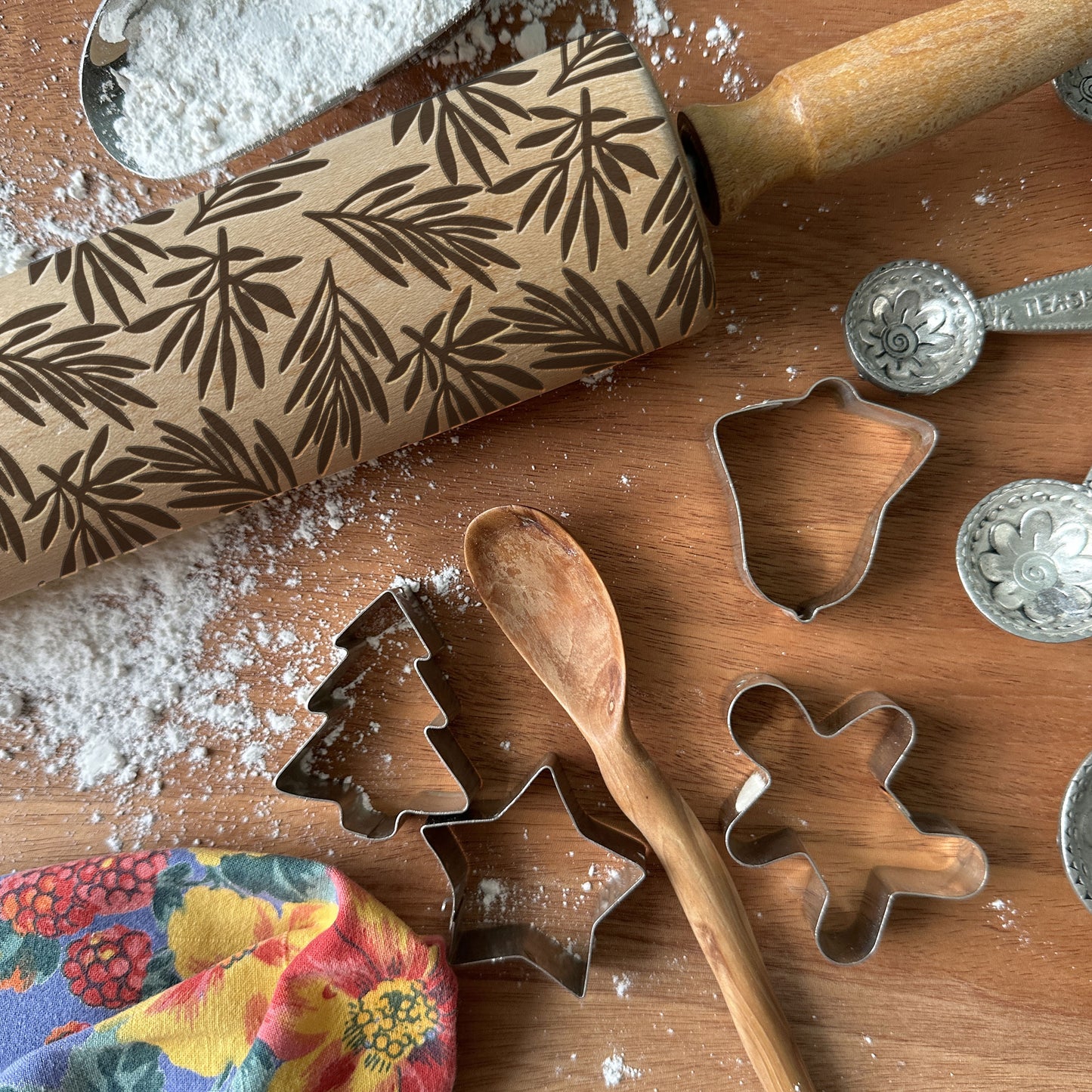 Baking tools including a patterned rolling pin, cookie cutters, and measuring spoons on a wooden surface with flour.