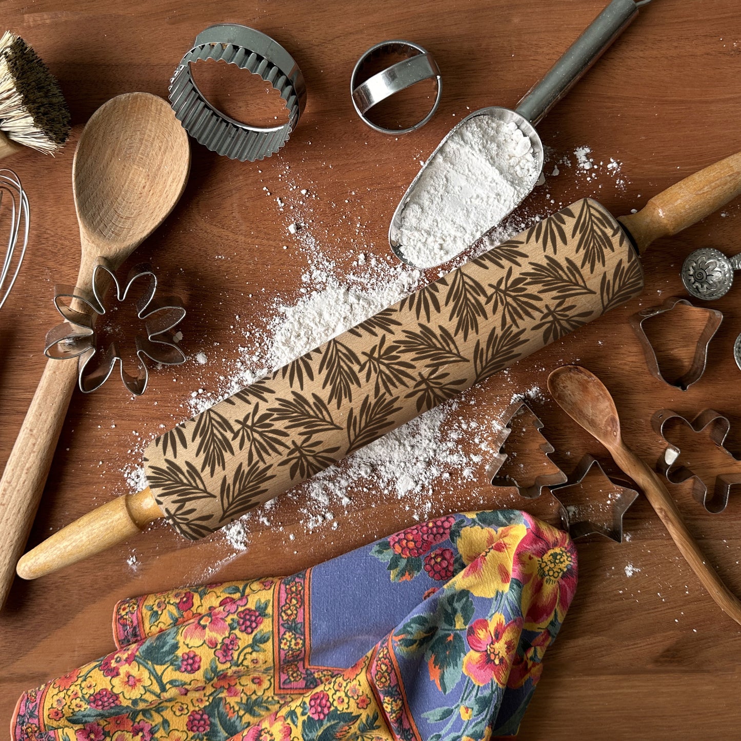 Wooden rolling pin with floral pattern on a wooden surface with flour and baking tools.