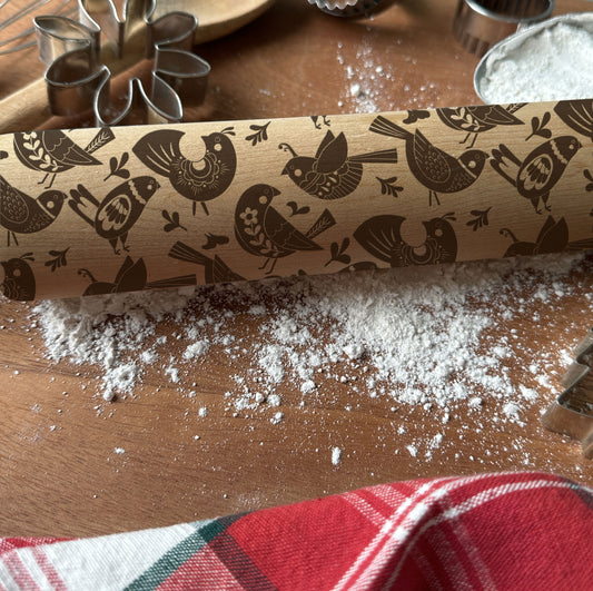 Rolling pin with bird pattern on a wooden surface with flour and cookie cutters.