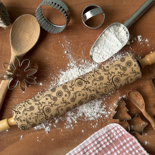 Baking tools including a rolling pin with floral patterns, wooden spoon, and cookie cutters on a wooden surface with flour.