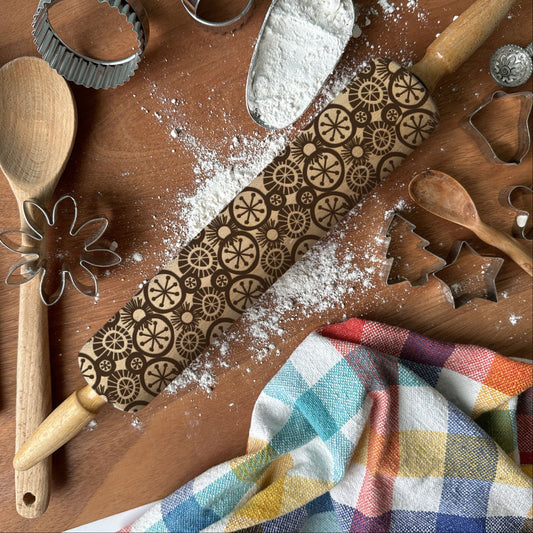 Baking tools including a rolling pin with embossed patterns, wooden spoon, whisk, and flour on a wooden surface.