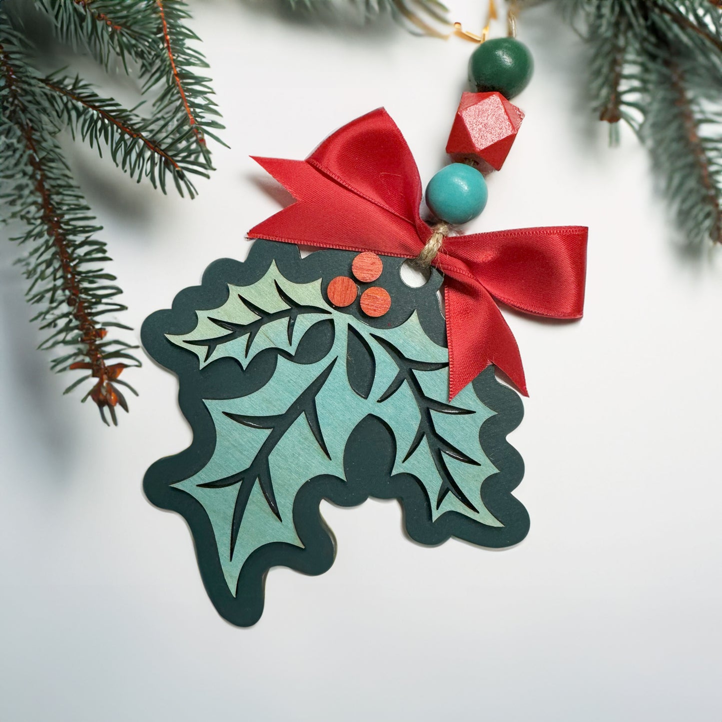 Decorative Christmas ornament with holly design, red bow, and beads on a white background.
