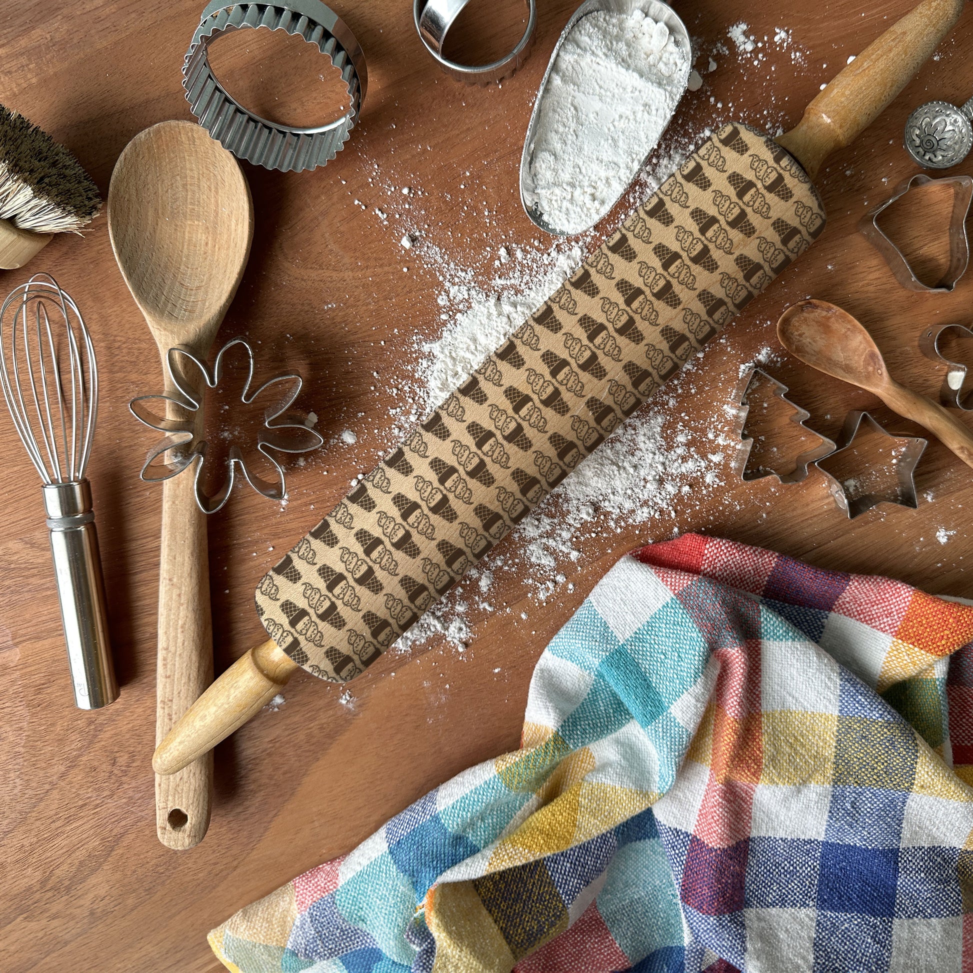 Baking tools including a rolling pin with embossed designs, wooden spoon, whisk, and cookie cutters on a wooden surface with flour.