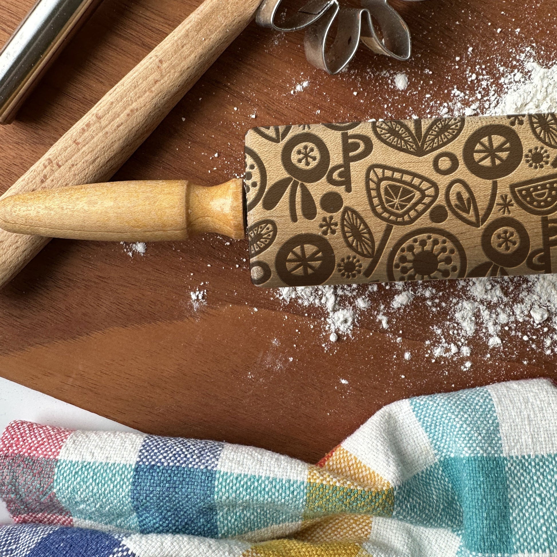 Decorative rolling pin with pattern on a wooden surface with flour and a checkered cloth.