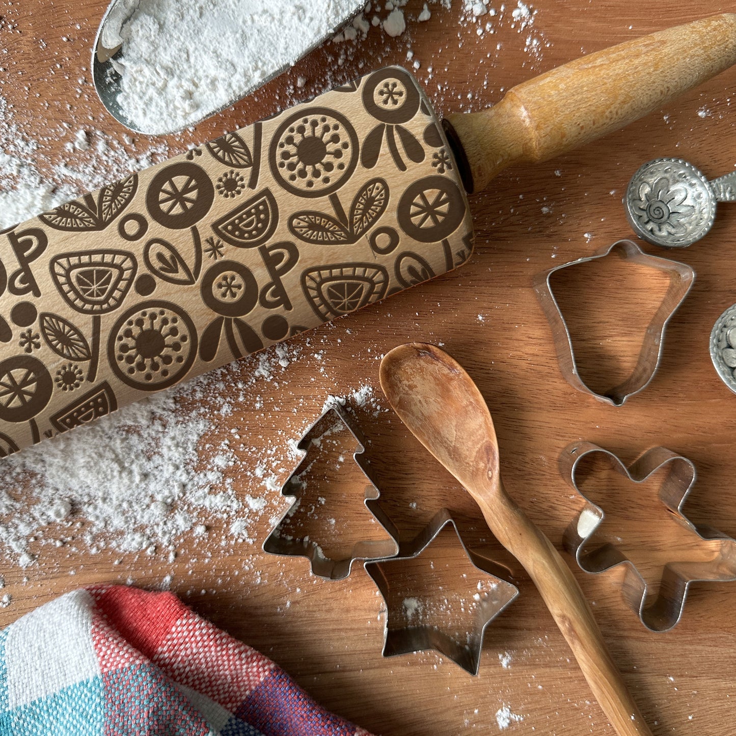 Baking tools including a patterned rolling pin, cookie cutters, and a wooden spoon on a wooden surface with flour.