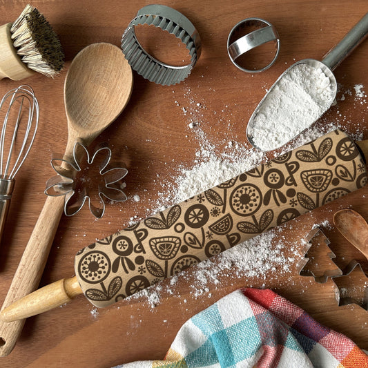 Baking tools including a rolling pin with decorative patterns, wooden spoon, whisk, and cookie cutters on a wooden surface.