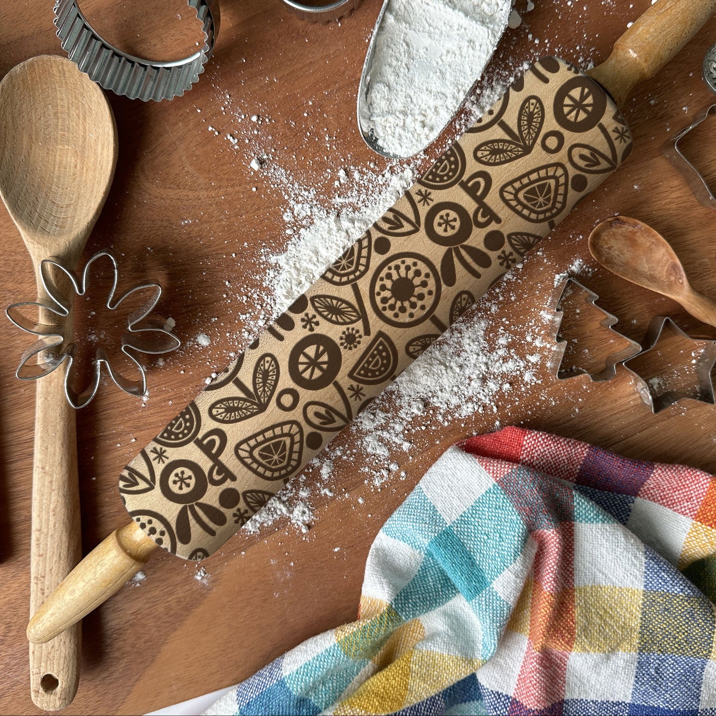 Baking tools including a rolling pin with patterns, wooden spoon, whisk, and cookie cutters on a wooden surface with flour.
