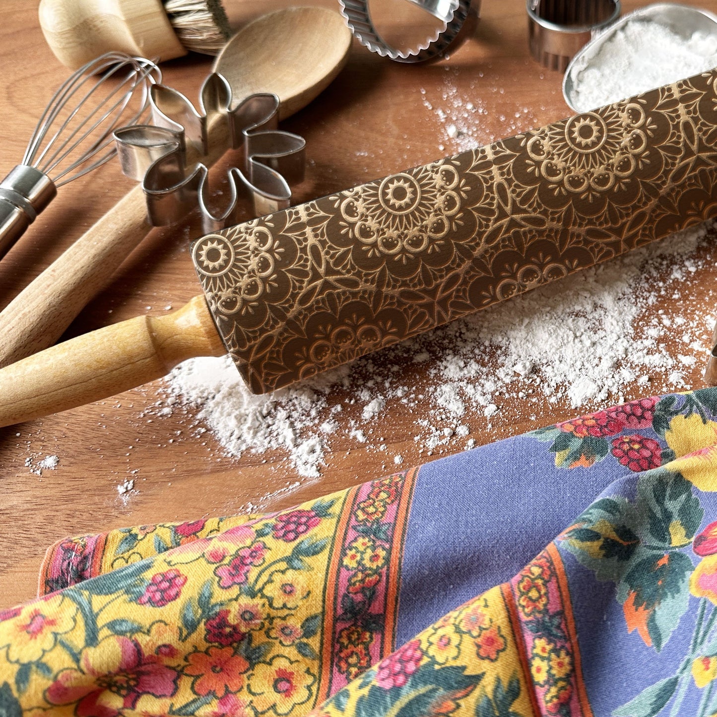 Decorative rolling pin with floral pattern on a wooden surface with baking tools and a colorful kitchen towel.