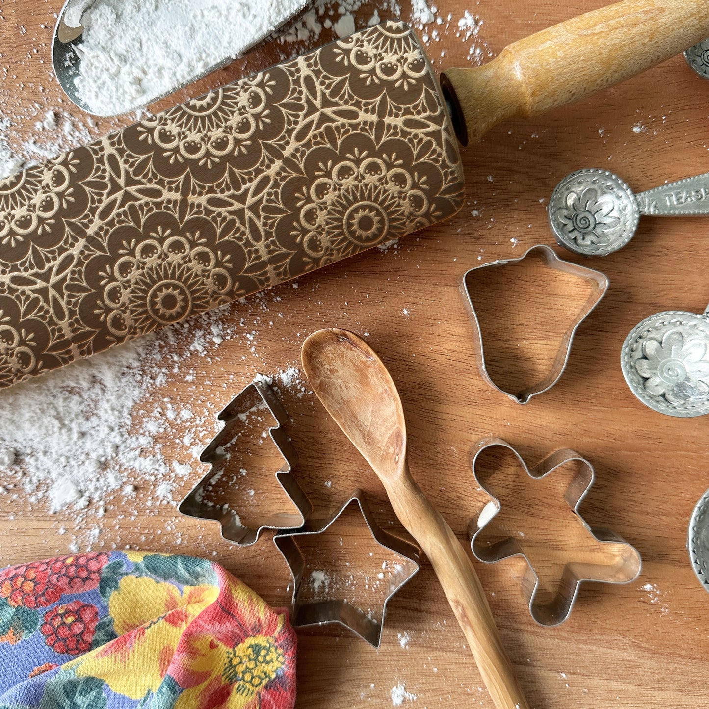 Baking tools including a patterned rolling pin, cookie cutters, and measuring spoons on a wooden surface with flour.