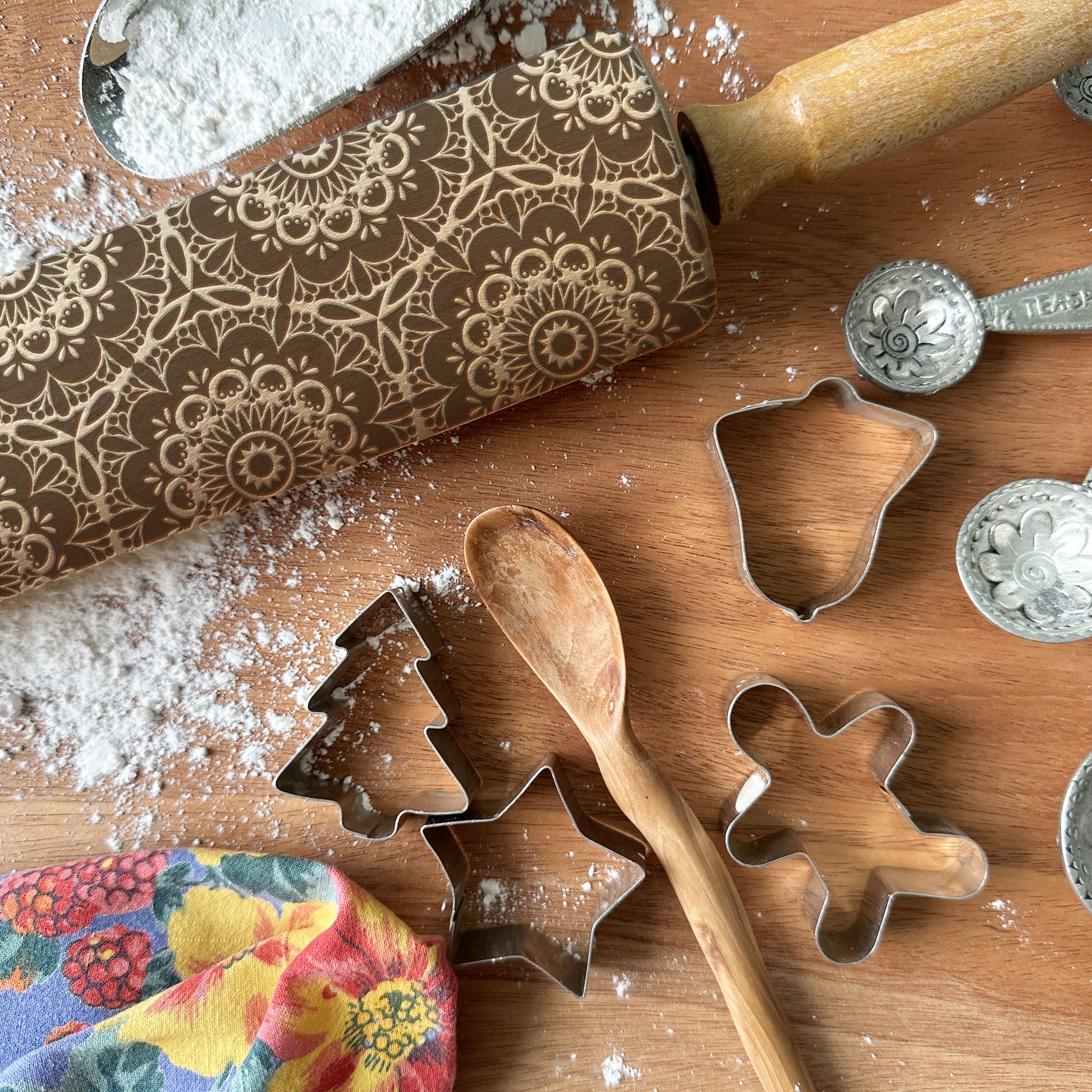 Baking tools including a patterned rolling pin, cookie cutters, and measuring spoons on a wooden surface with flour.