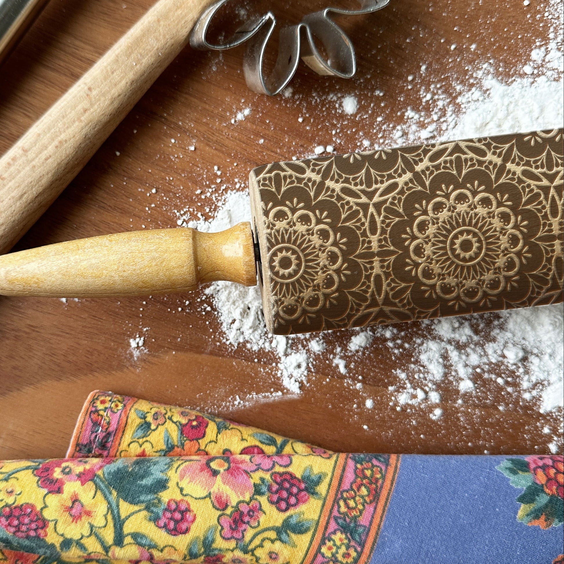 Decorative rolling pin with floral patterns on a wooden surface with flour and cookie cutters.