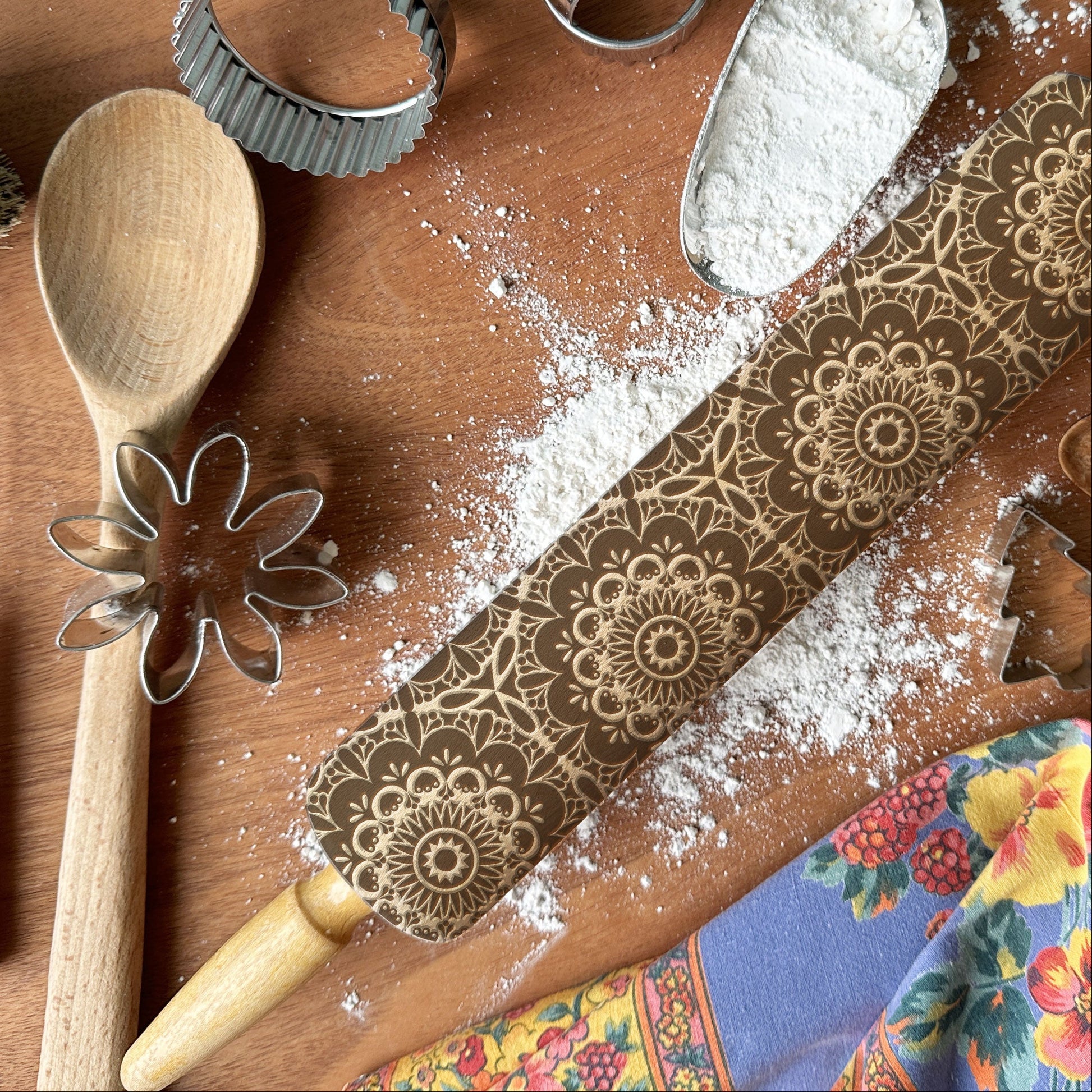 Baking tools including a wooden spoon, rolling pin with floral patterns, and cookie cutters on a wooden surface.