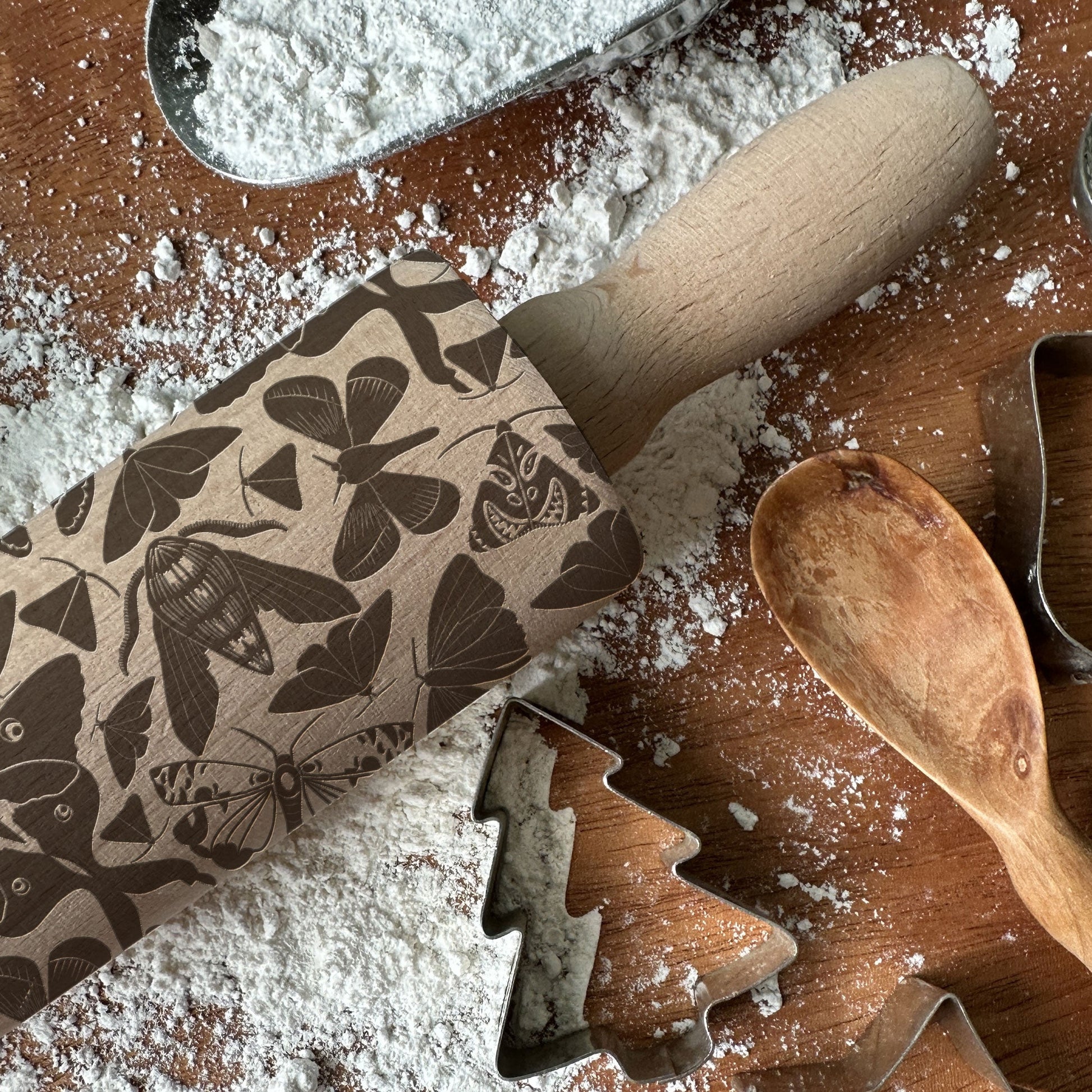 Butterfly-patterned rolling pin on a flour-dusted surface with wooden spoon and cookie cutters.