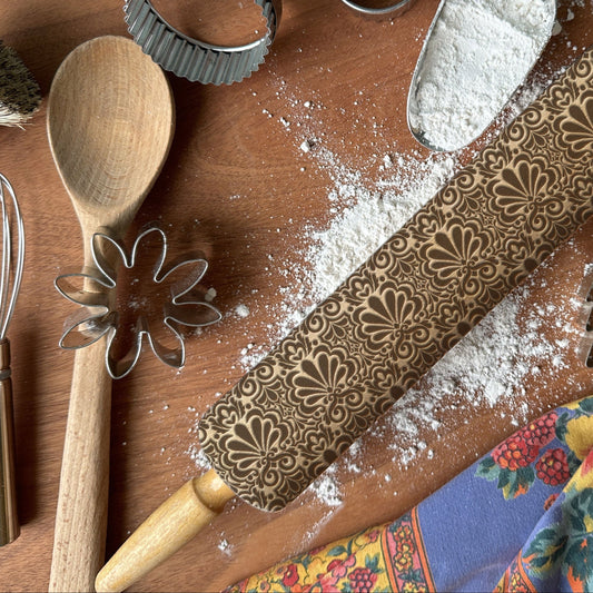 Baking tools including a wooden spoon, rolling pin with floral patterns, and flour on a wooden surface.