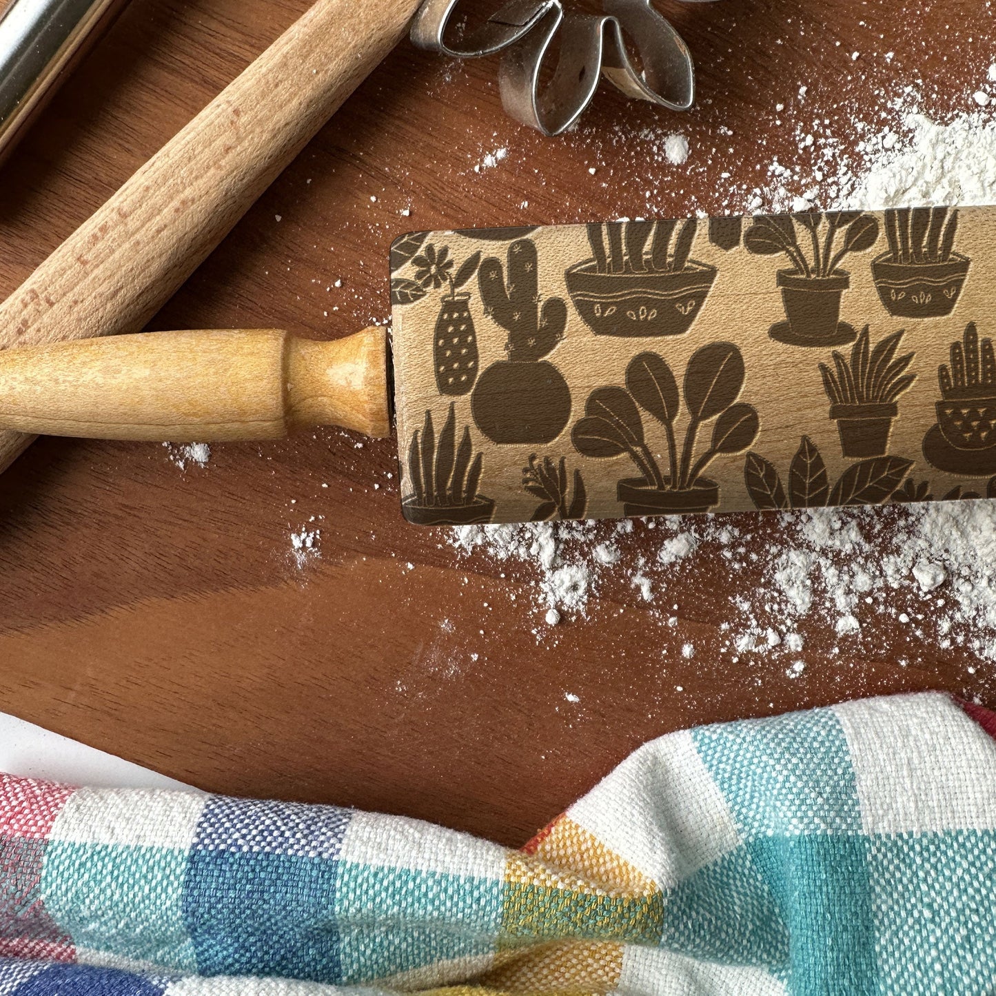 Rolling pin with cactus pattern on a wooden surface with flour and a checkered cloth.
