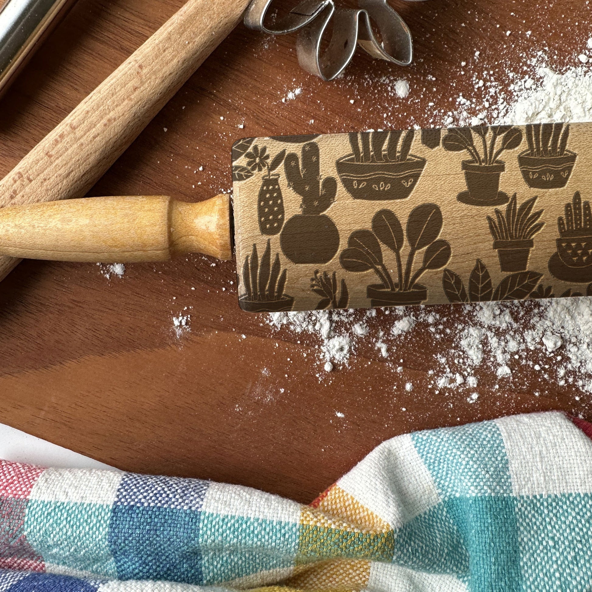 Rolling pin with cactus pattern on a wooden surface with flour and a checkered cloth.