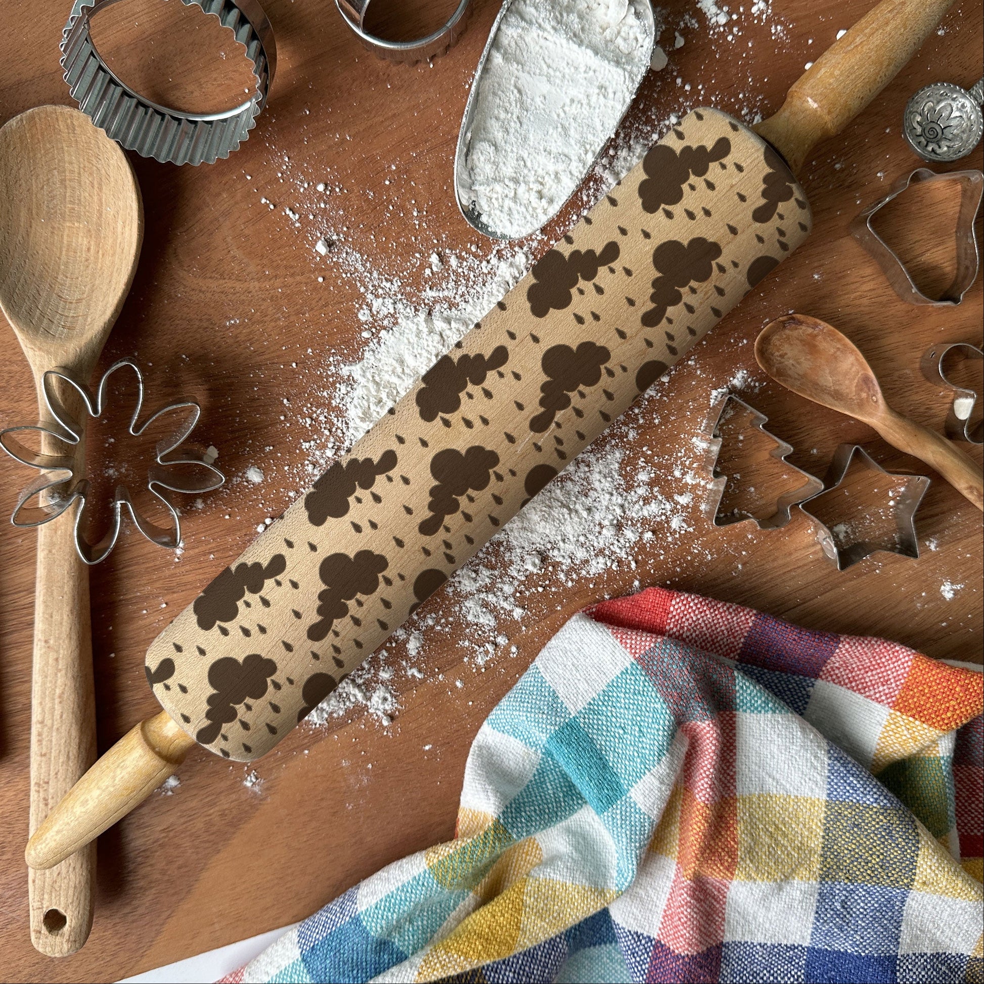 Baking tools including a rolling pin with cookie stamps, wooden spoon, whisk, and flour on a wooden surface.
