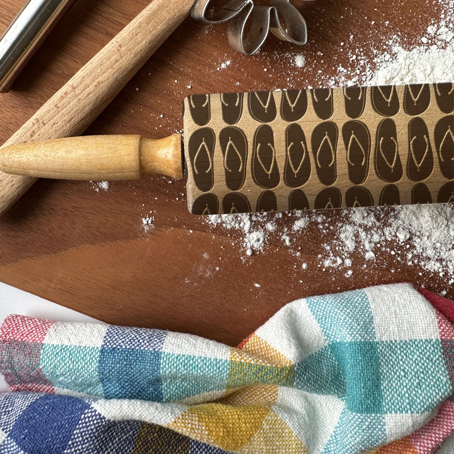 Wooden rolling pin with embossed designs on a wooden surface with flour and a checkered cloth.