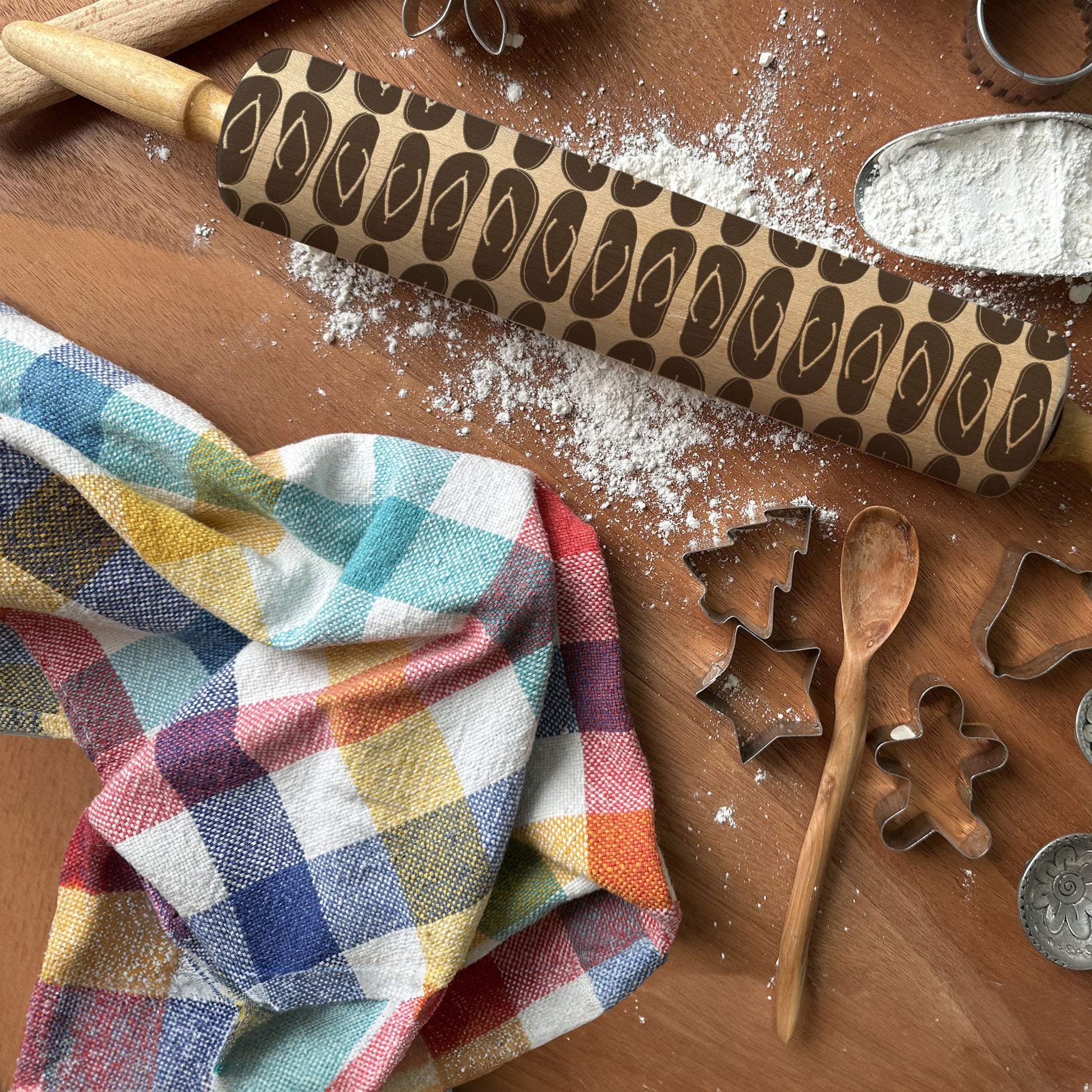 Wooden rolling pin with decorative pattern on a wooden surface with cookie cutters and a colorful towel.