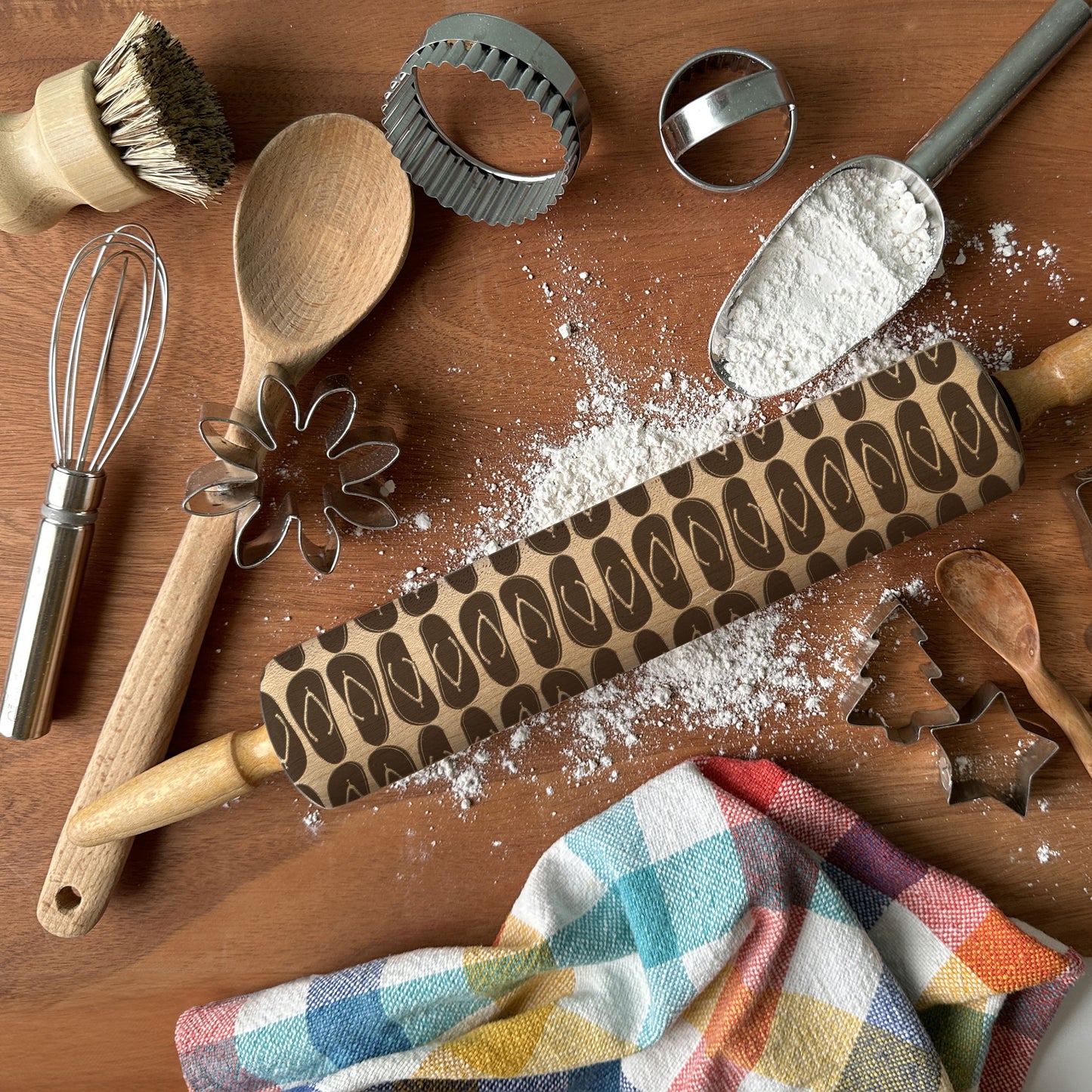 Baking tools including a rolling pin with a pattern, wooden spoon, whisk, and cookie cutters on a wooden surface.